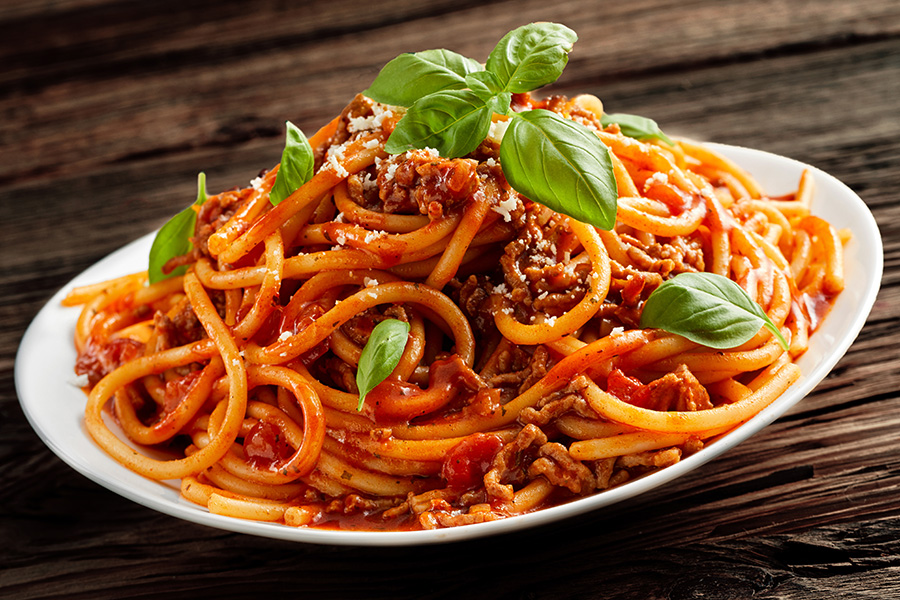 A plate of spaghetti with meat sauce, garnished with fresh basil leaves and grated cheese, served on a white dish against a rustic wooden background.