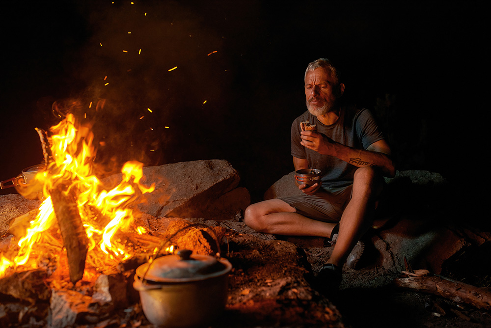 A man sits on rocky ground by a campfire at night, holding a cup and bowl, with a pot cooking over the flames. He appears relaxed, surrounded by darkness and illuminated by the firelight.